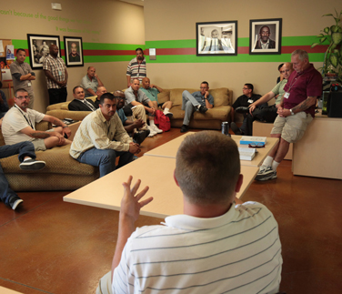 A group of men gathered in a casual meeting room, sitting on couches and chairs while one person speaks at the front. The room has framed portraits on the wall and a relaxed, community-centered atmosphere.