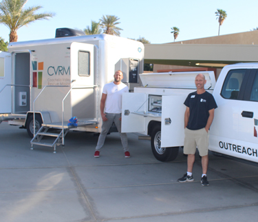 Two men standing beside a white outreach vehicle and a CVRM trailer equipped for mobile services. Both are smiling under the sun, with palm trees and buildings in the background, representing community outreach efforts.