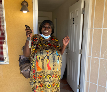 A joyful woman standing at the doorway of an apartment, smiling broadly while holding her phone and keys. She wears a colorful patterned dress and has a bag over her shoulder, with the door labeled “5” open behind her.