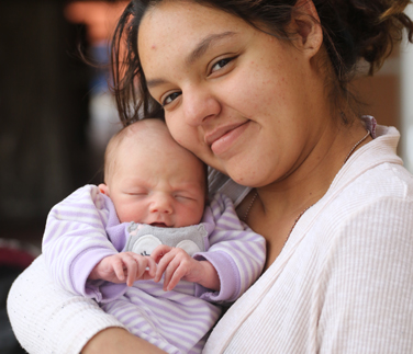 A mother smiling gently while holding her newborn baby dressed in soft lavender stripes. The baby sleeps peacefully against her chest, creating a tender and heartwarming moment.