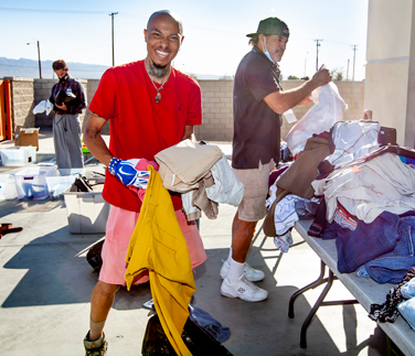 Smiling volunteer in a red shirt holding folded clothes while helping organize a clothing drive outdoors. Tables behind him are piled with garments, and other volunteers are sorting donations under bright sunlight.