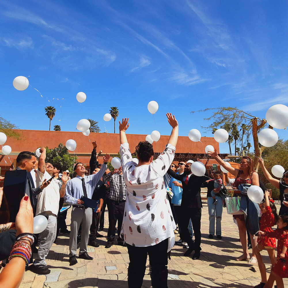 A joyful outdoor celebration with people releasing white balloons into a clear blue sky. The central person has their arms raised, surrounded by smiling participants, palm trees, and warm sunlight.