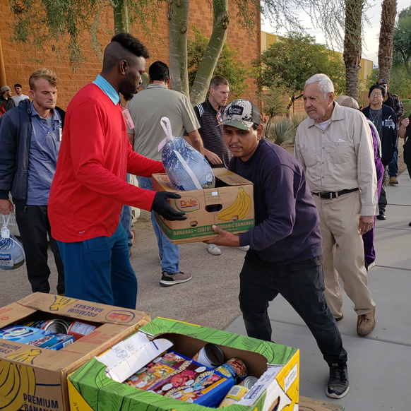 Smiling volunteer in a red shirt holding folded clothes while helping organize a clothing drive outdoors. Tables behind him are piled with garments, and other volunteers are sorting donations under bright sunlight.