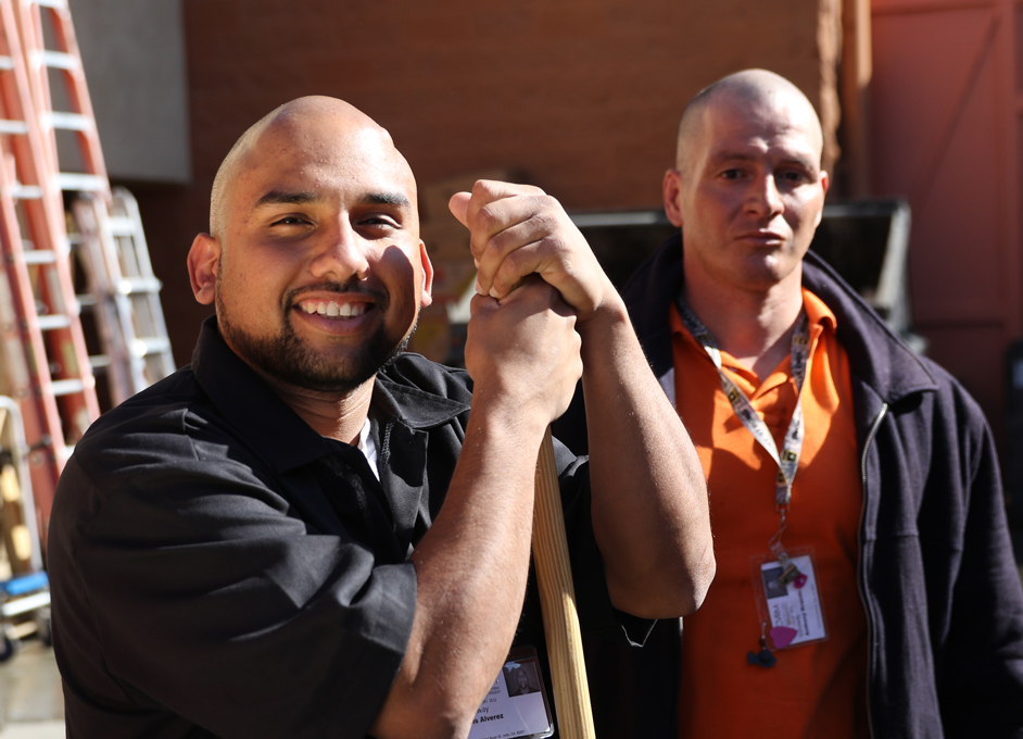 Two men standing outdoors, one smiling and holding a broom handle, both wearing work badges and casual uniforms with ladders and a building in the background.