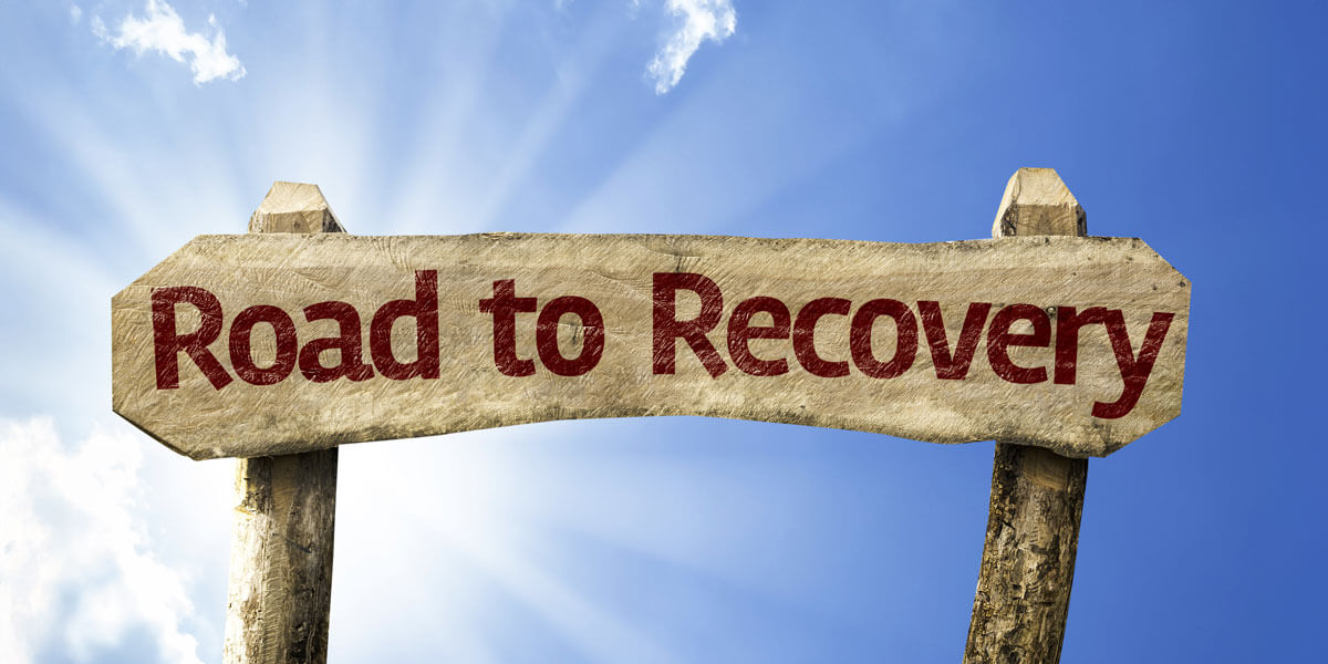 A wooden sign reading “Road to Recovery” stands against a bright blue sky with sunlight shining behind it, symbolizing hope and healing.