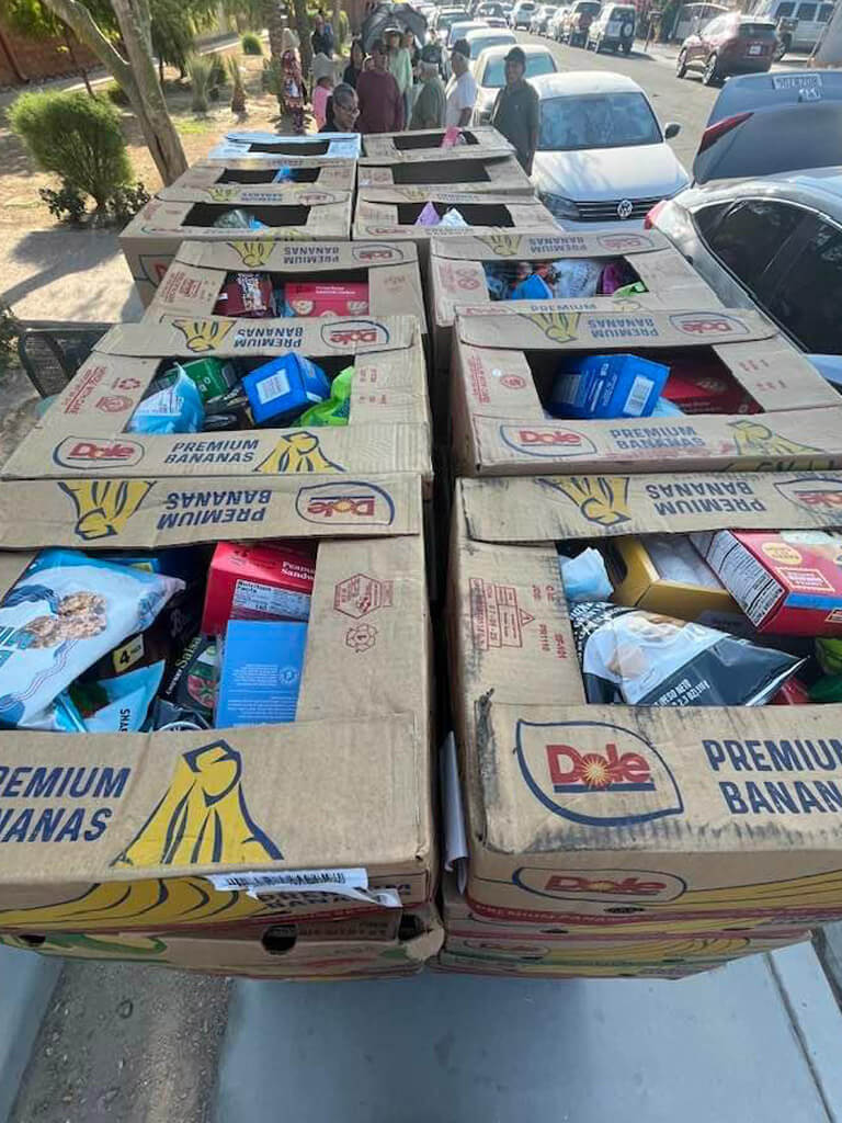 Cardboard boxes filled with packaged food items are stacked in the back of a vehicle, with people waiting nearby as part of a community food distribution effort.