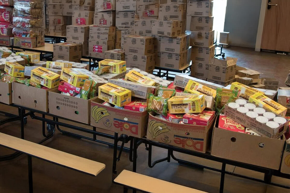 Long tables lined with boxes of groceries and canned goods inside a large room, prepared for organized community food distribution.