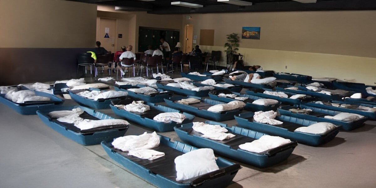 Rows of temporary beds are arranged inside a large shelter space while people gather in the background, highlighting emergency housing support services.