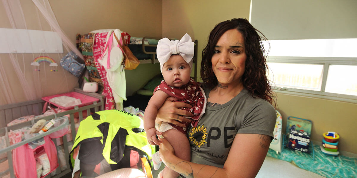 A woman wearing a gray 'Hope' t-shirt holds her infant daughter, who has a large white bow headband, while standing in a small shelter room filled with a crib, bunk bed, baby supplies, and colorful belongings.