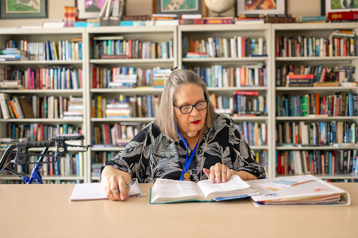 An older woman with gray hair and glasses sits at a table reading an open book in front of floor-to-ceiling bookshelves, with a walker visible at her side.