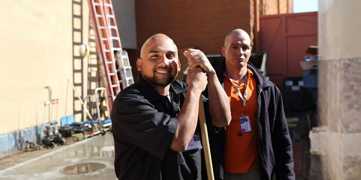 Two men wearing lanyards share a friendly handshake while standing together outdoors at a worksite with ladders and equipment in the background.