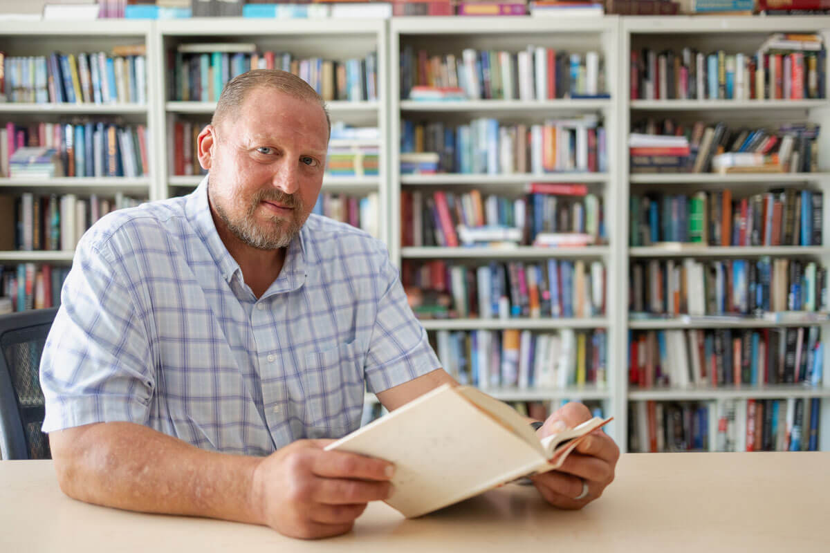 A smiling man with a beard sits at a desk holding an open book in front of packed bookshelves.