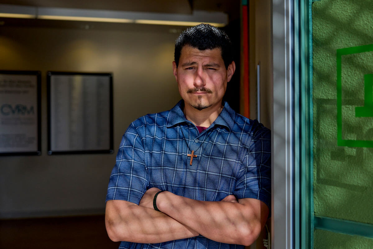 A man with a goatee, wearing a blue plaid short-sleeved shirt and a wooden cross necklace, stands with his arms crossed while leaning against a sunlit doorframe.