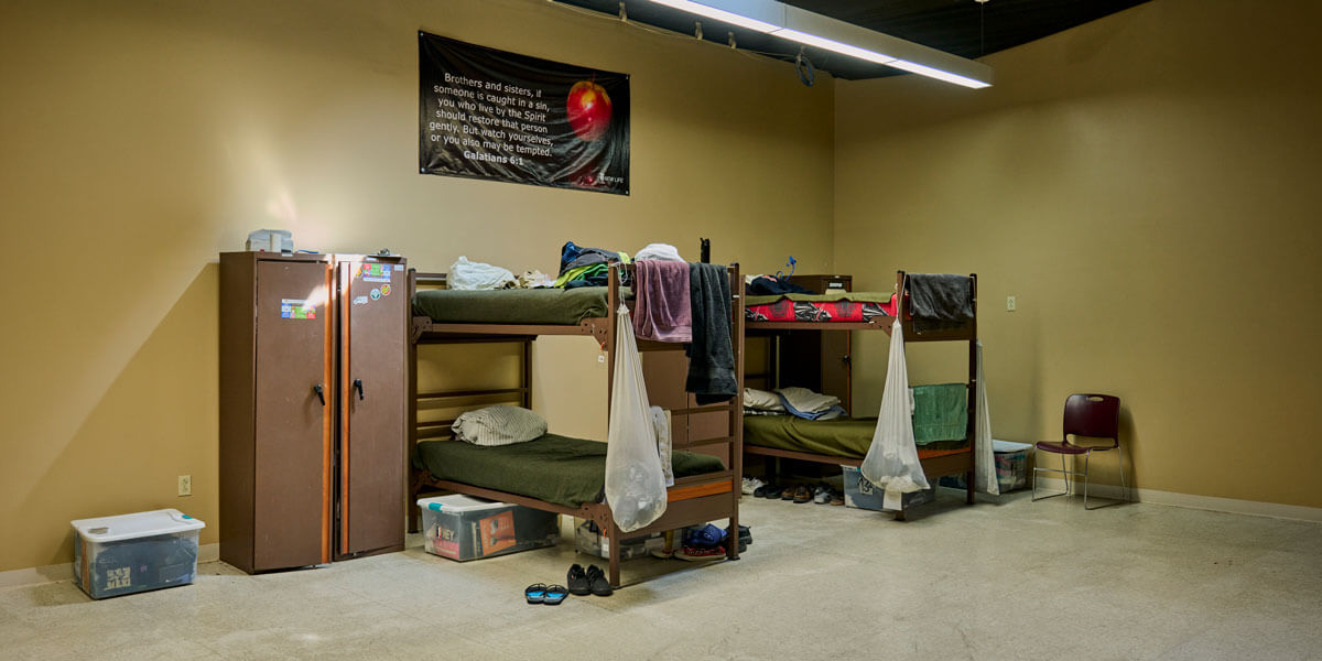 A sparse dormitory room contains two metal bunk beds with scattered belongings, a brown metal locker, clear storage bins on the floor, and a wall banner displaying a Bible verse.