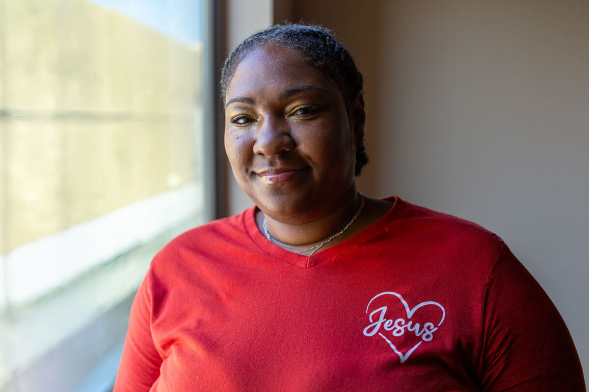 Woman in a red shirt with a heart shaped Jesus graphic smiling gently near a window.