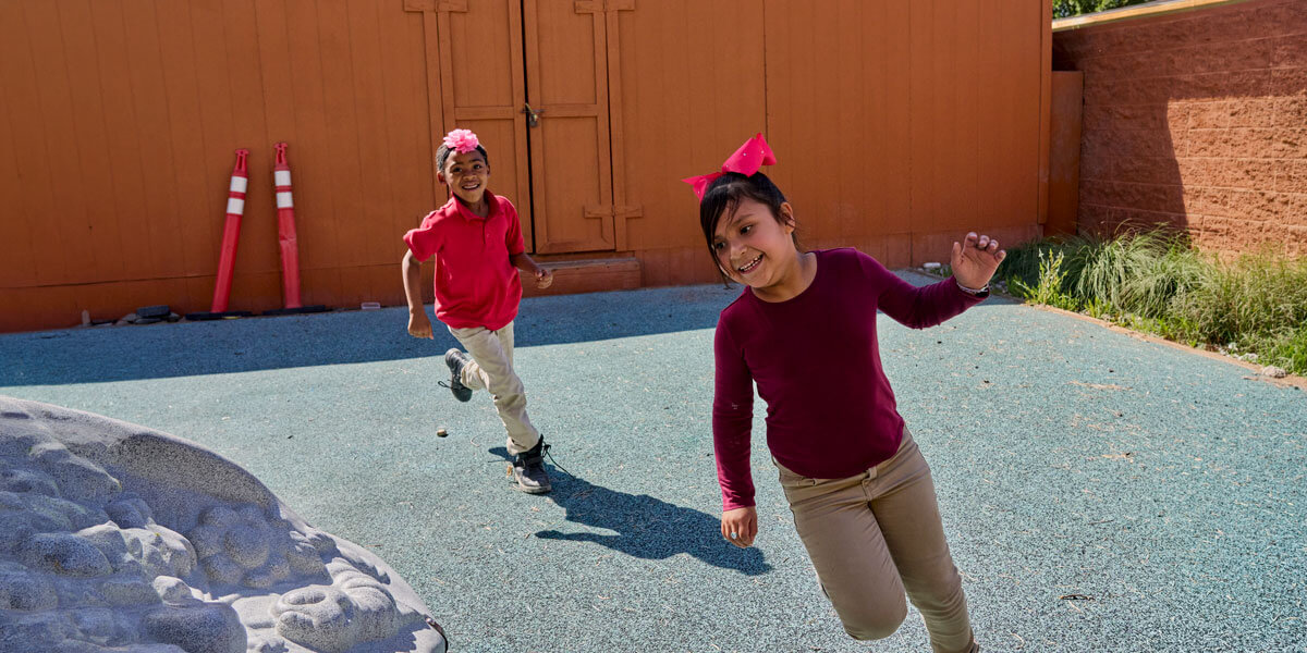 Two smiling children in school uniforms running across a playground on a sunny day.