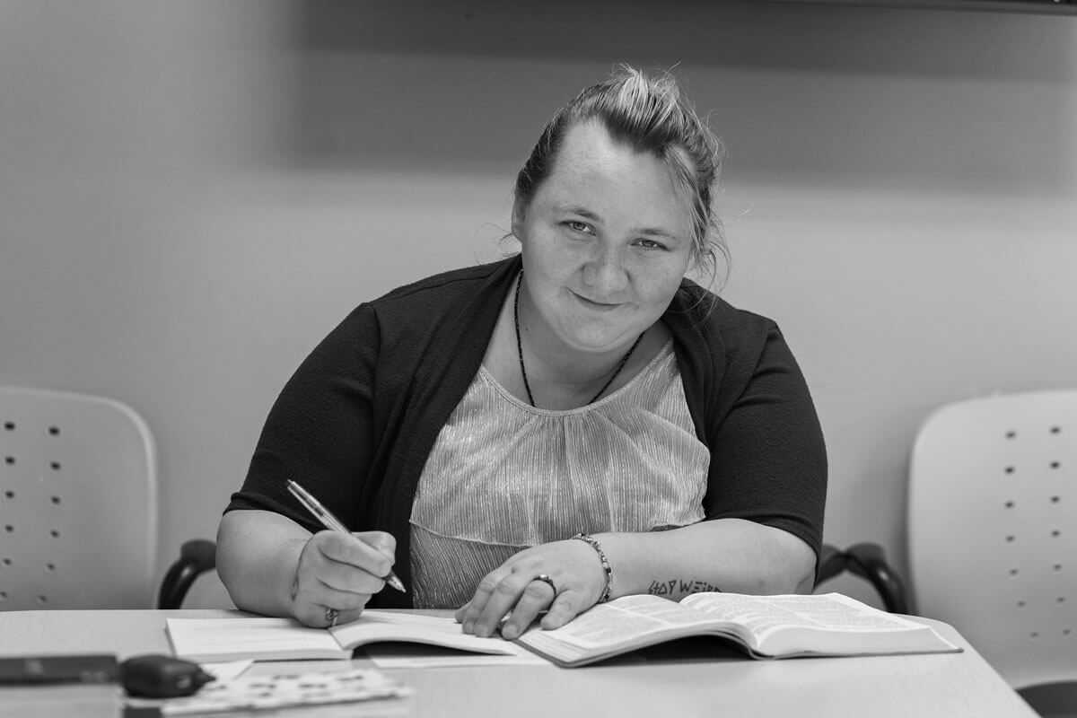 A smiling woman sits at a table writing in a notebook with an open book in front of her.