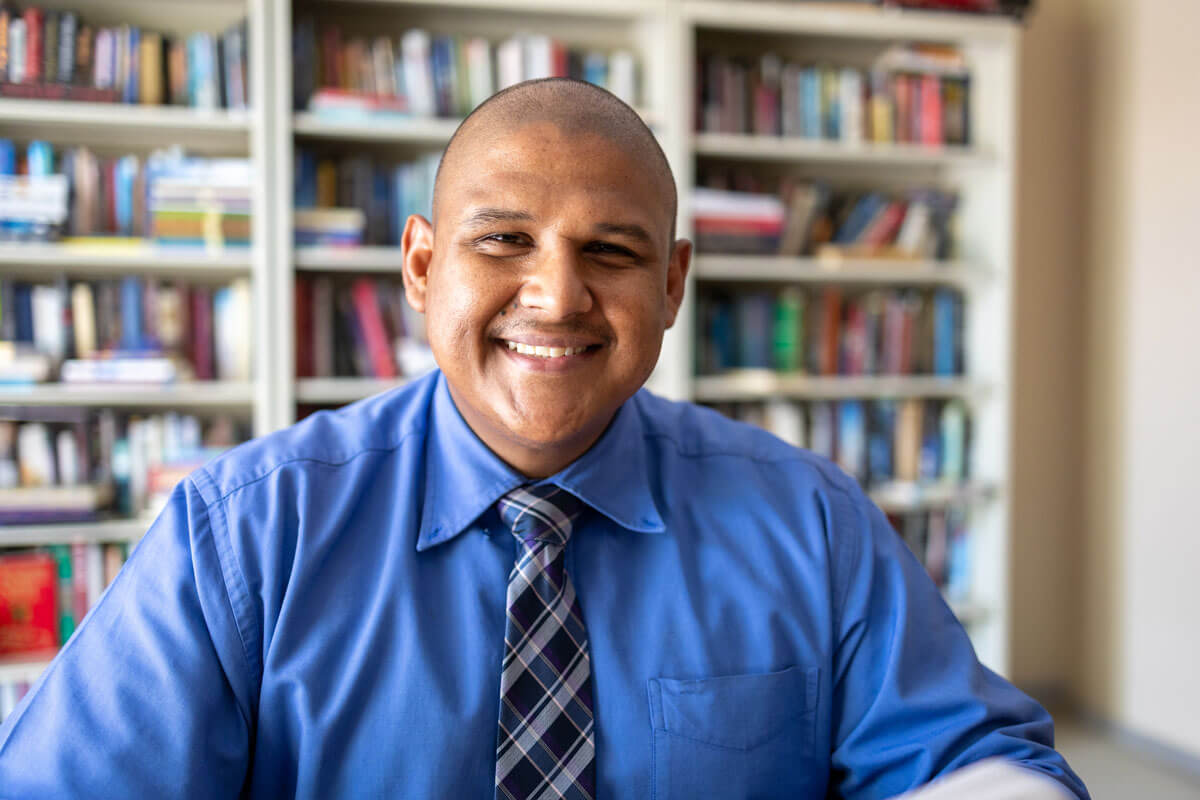 A smiling man in a blue collared shirt and plaid tie sits in front of full bookshelves.